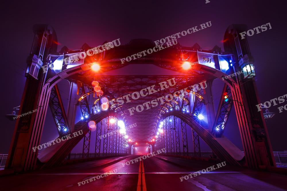 На фотографии изображён Большеохтинский мост в Санкт-Петербурге, который подсвечен в ночное время. Его уникальная архитектура и подсветка делают его заметной ориентировкой в городском ночном пейзаже.